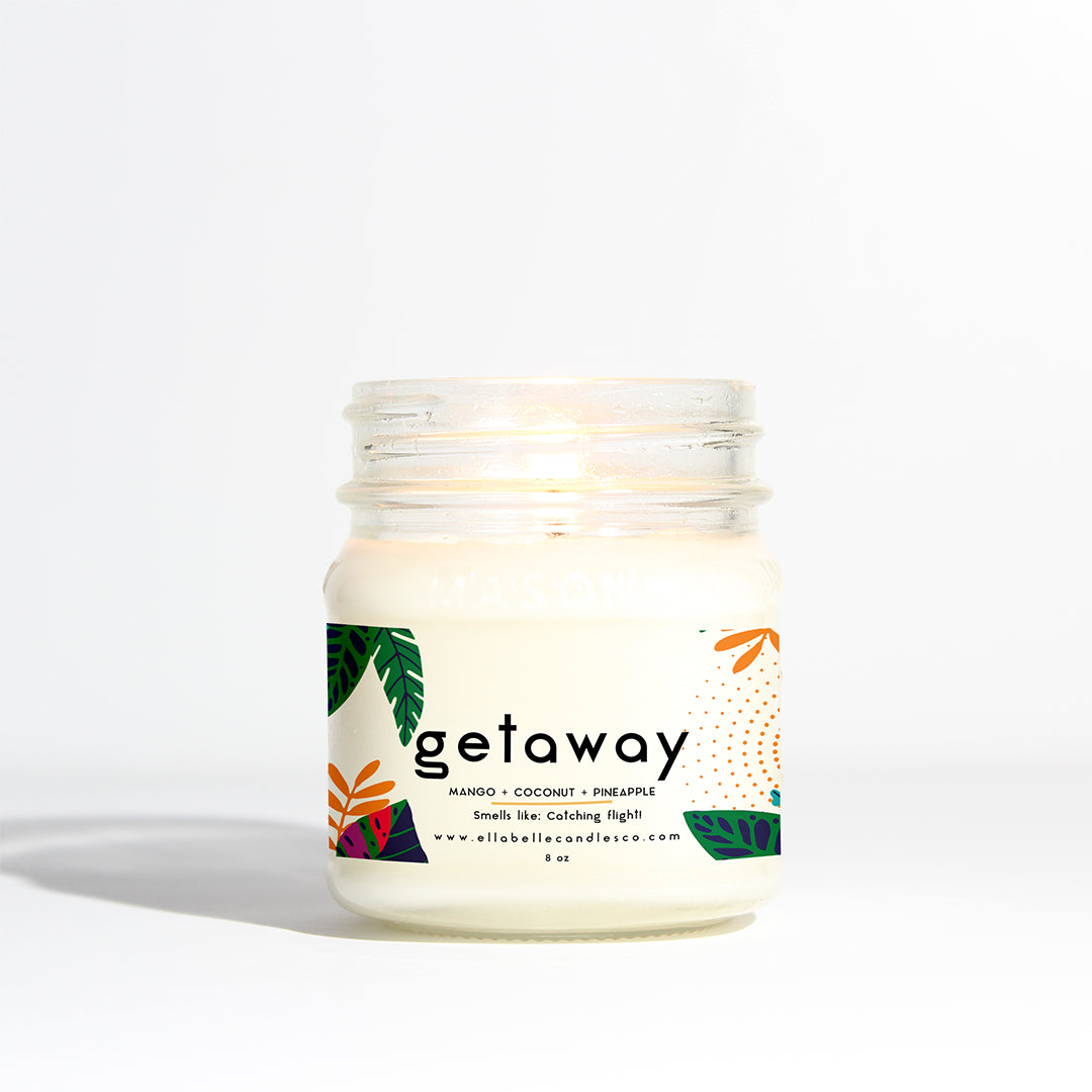 A mason jar candle with a white label that has the product name 'Getaway' and a brief description. The candle is lit and has a tropical design with coconut, pineapple, and mango illustrated on the label.