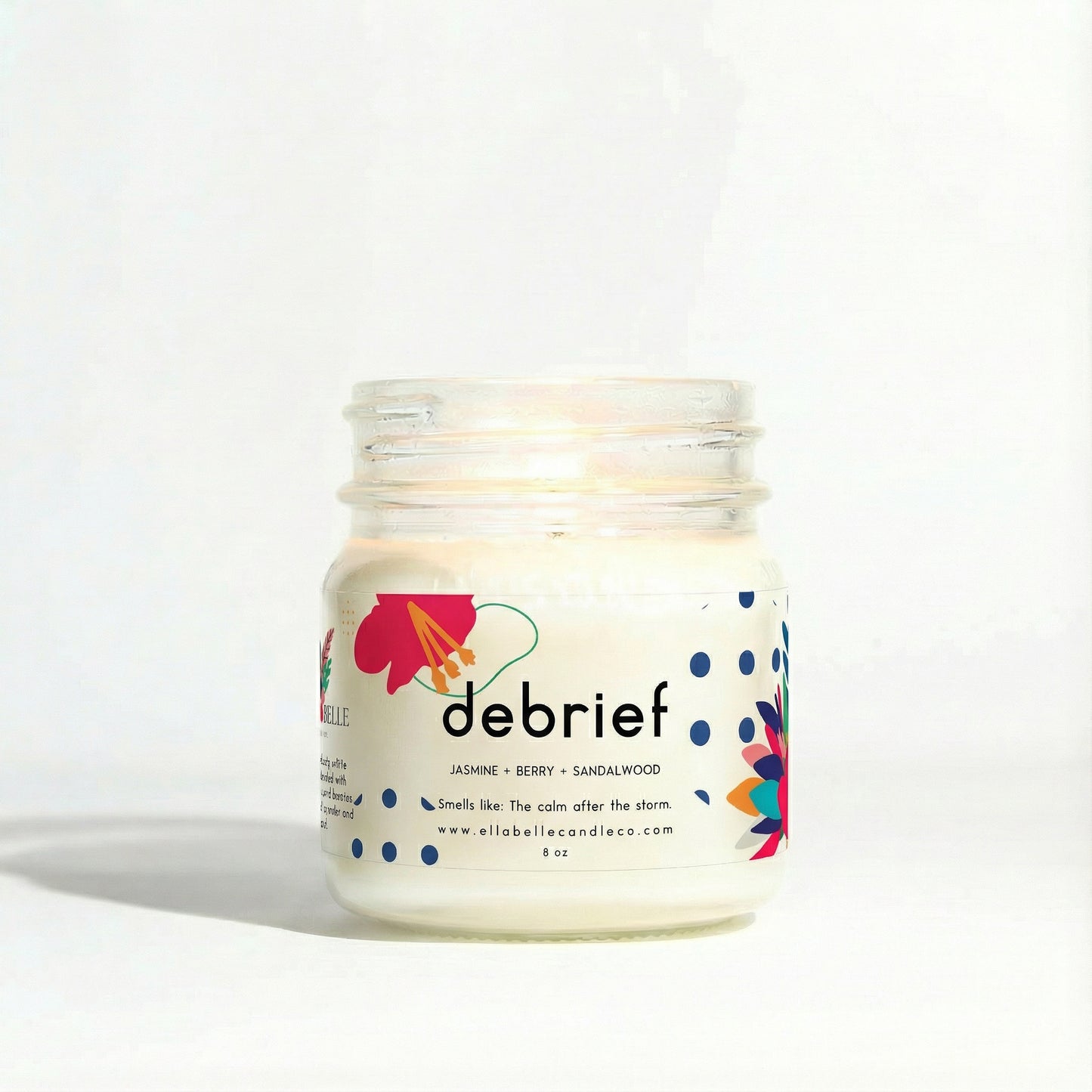 A clear mason jar candle with a label that reads 'DEBRIEF' and has a colorful flower design. The candle is lit and has a white wax base