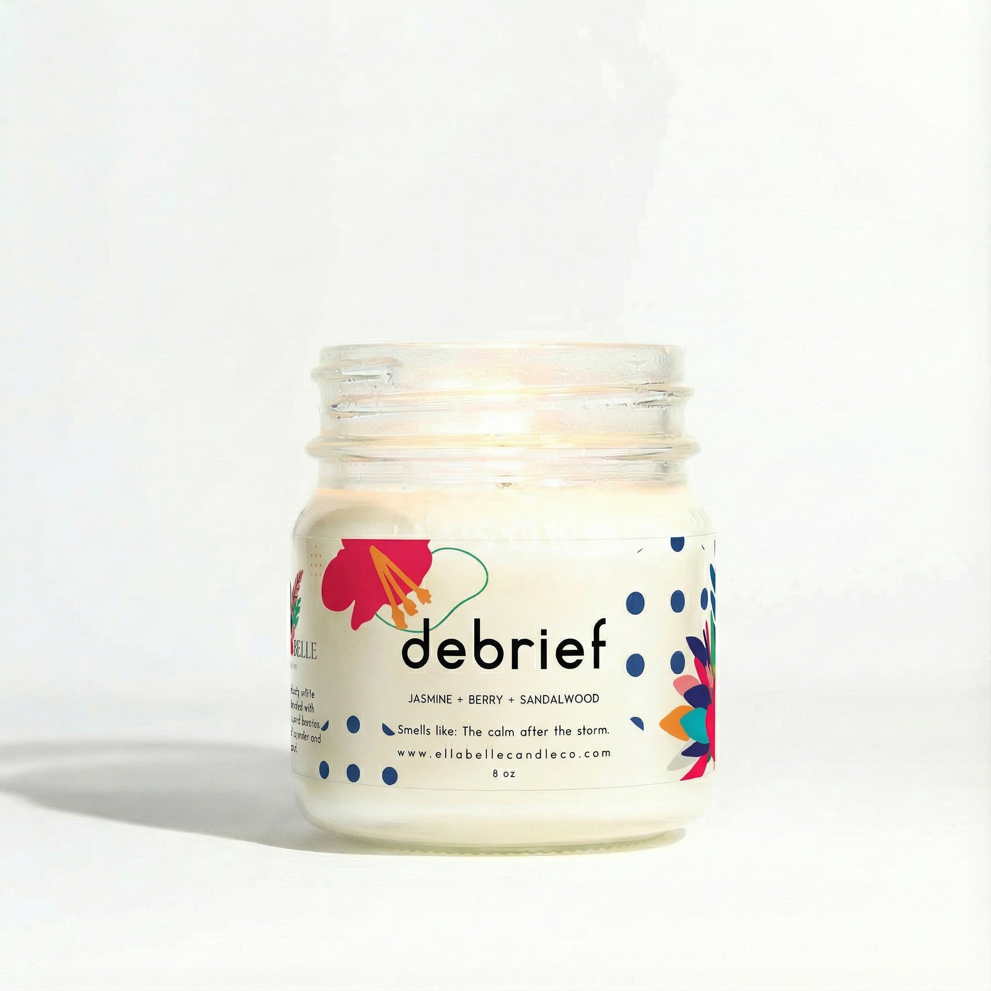A clear mason jar candle with a label that reads 'DEBRIEF' and has a colorful flower design. The candle is lit and has a white wax base