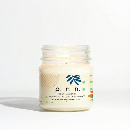 A mason jar candle with a label that reads 'P.R.N.', coconut + lemongrass with a white wax cap and a lit wick.