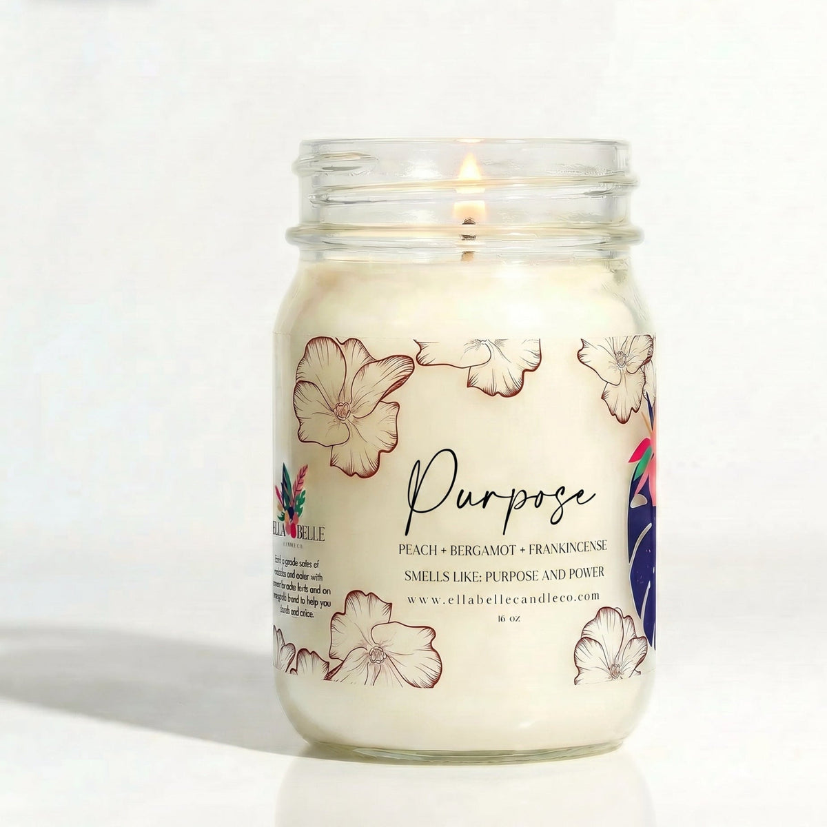 A lit white candle in a clear glass mason jar with floral illustrations and the word 'Purpose' on the label, photographed against a plain white background. The brand name 'Ella Belle Candle Co' is visible on the label.