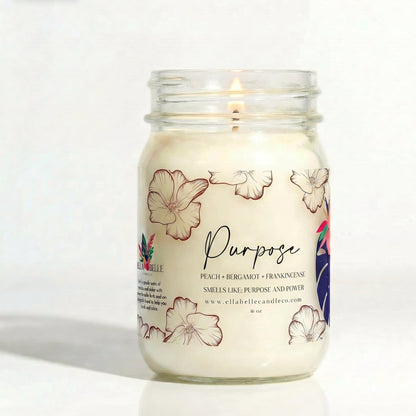 A lit white candle in a clear glass mason jar with floral illustrations and the word 'Purpose' on the label, photographed against a plain white background. The brand name 'Ella Belle Candle Co' is visible on the label.