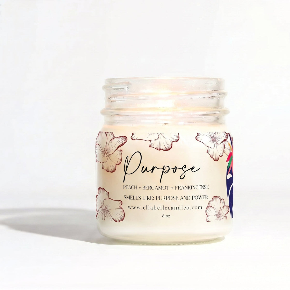 A lit jar candle with a clear glass body and white wax, featuring a label with peach floral illustrations and the word 'Purpose' in script. The candle is photographed on a clean white background with a soft shadow.