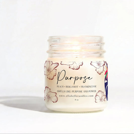 A lit jar candle with a clear glass body and white wax, featuring a label with peach floral illustrations and the word 'Purpose' in script. The candle is photographed on a clean white background with a soft shadow.