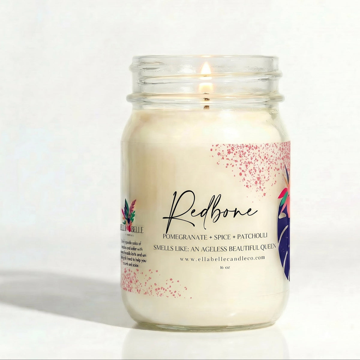 A white candle in a clear glass jar with a printed label reading 'Redbone' and 'Pomegranate + Spice + Patchouli,' photographed on a plain white background. 