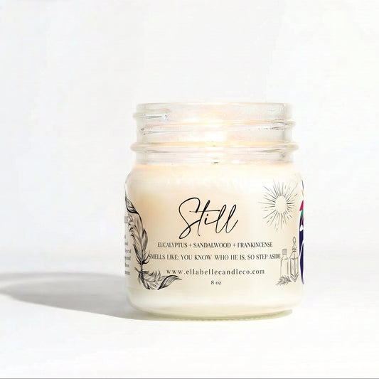A lit jar candle in a clear glass mason jar with cream-colored wax and a white label reading 'Still' and 'Eucalyptus + Sandalwood + Frankincense'. The brand name 'ella belle candle co.