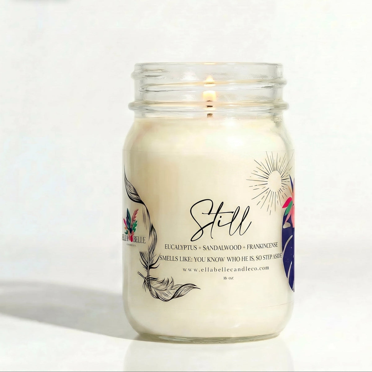 A lit white soy wax candle in a clear glass mason jar with a white label reading 'Still' and feather illustrations, set against a plain white background.


