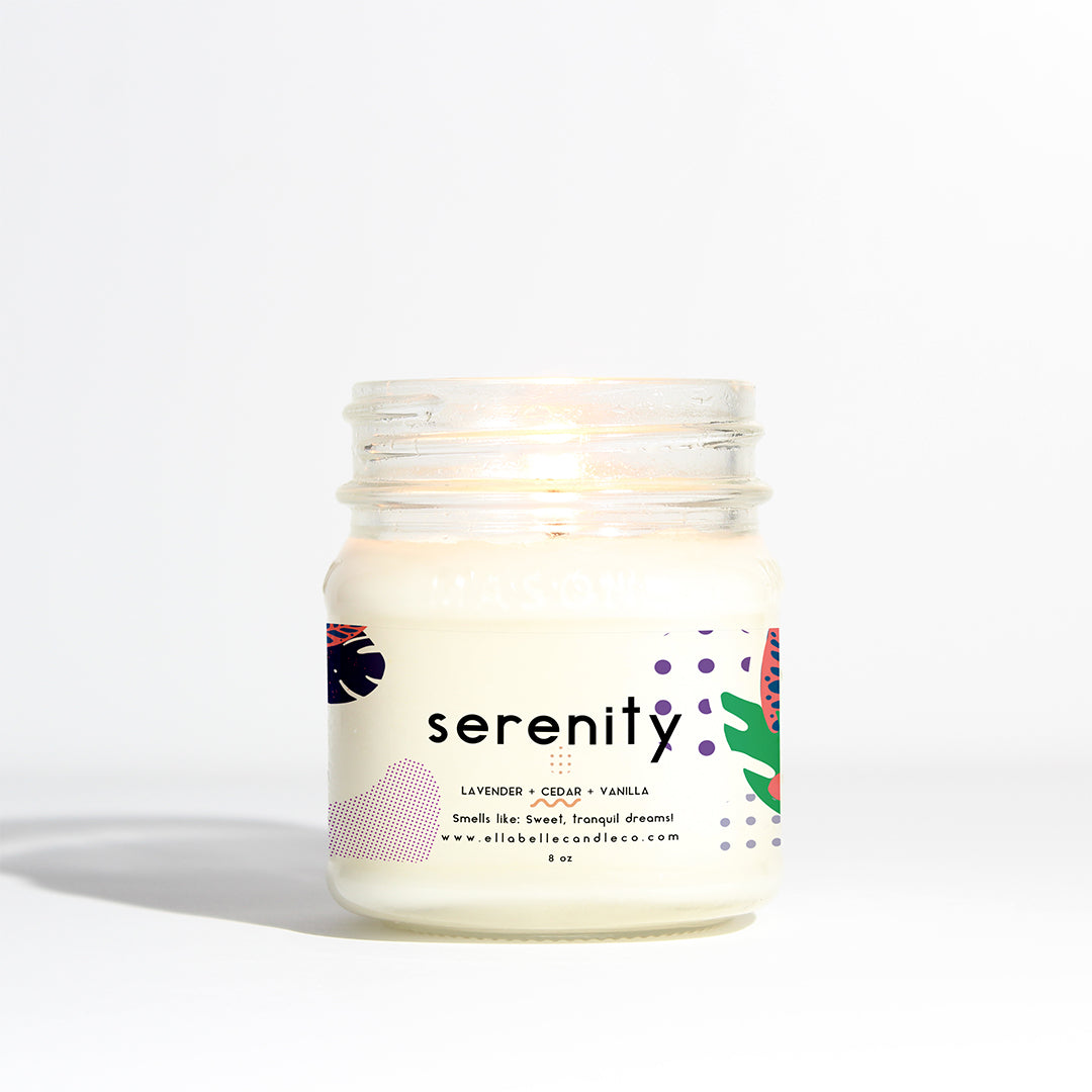 A clear mason jar candle with a label that reads 'Serenity' and lists the scents as lavender, cedar, violet, musk, and vanilla with a dash of rose. The candle is lit, and the label also provides the brand's website.