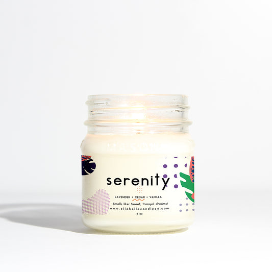 A clear mason jar candle with a label that reads 'Serenity' and lists the scents as lavender, cedar, violet, musk, and vanilla with a dash of rose. The candle is lit, and the label also provides the brand's website.